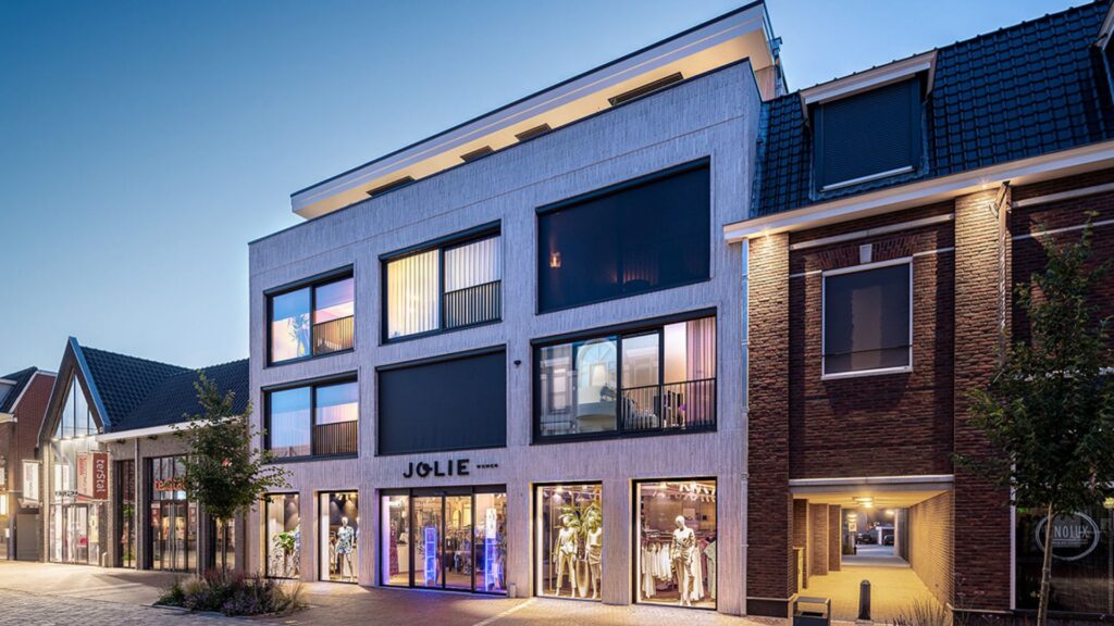 Een moderne winkelgevel met bovenliggende appartementen. Op de begane grond bevindt zich de kledingwinkel "Jolie Women". Het gebouw heeft grote ramen en grijze rolluiken, en is verlicht in de avond. Links en rechts van het gebouw zijn andere winkels zichtbaar. | Brick Finesse